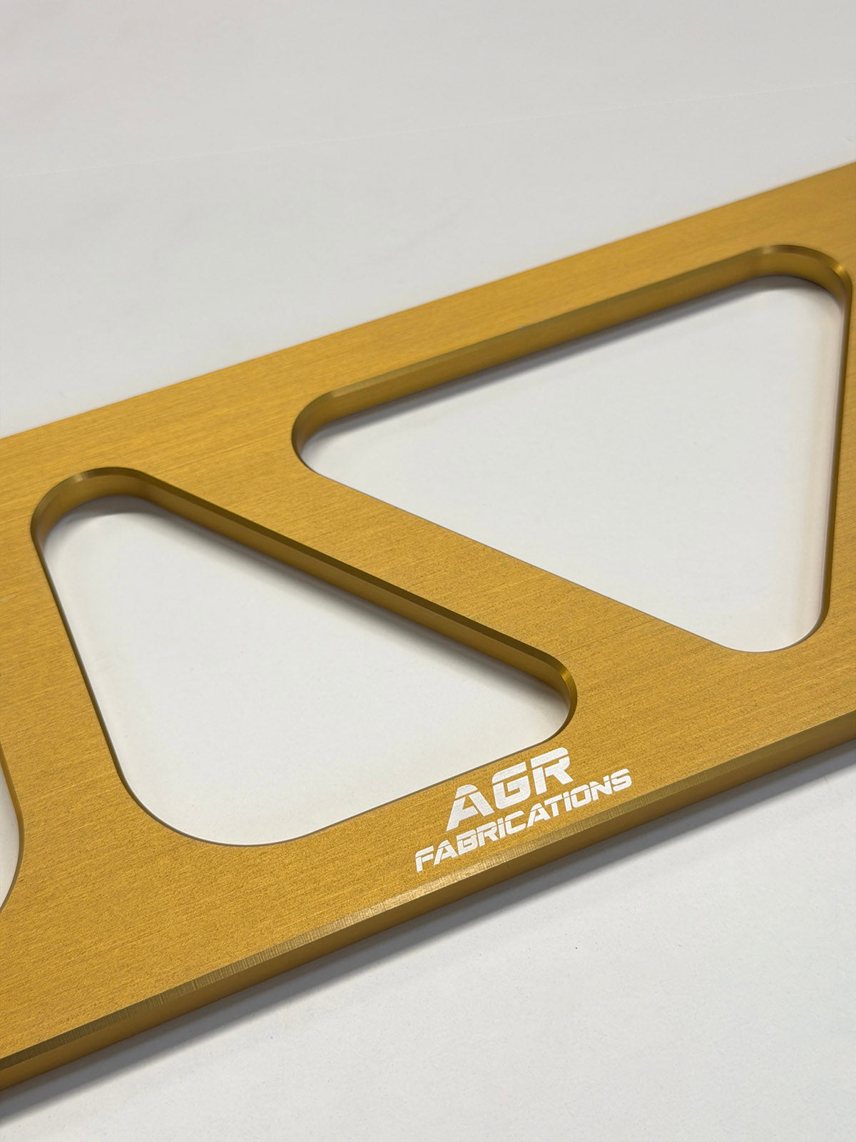 A close-up of a gold anodized metal bracket or frame piece with triangular cutouts and the white engraved text 'AGR FABRICATIONS' on a light gray background.